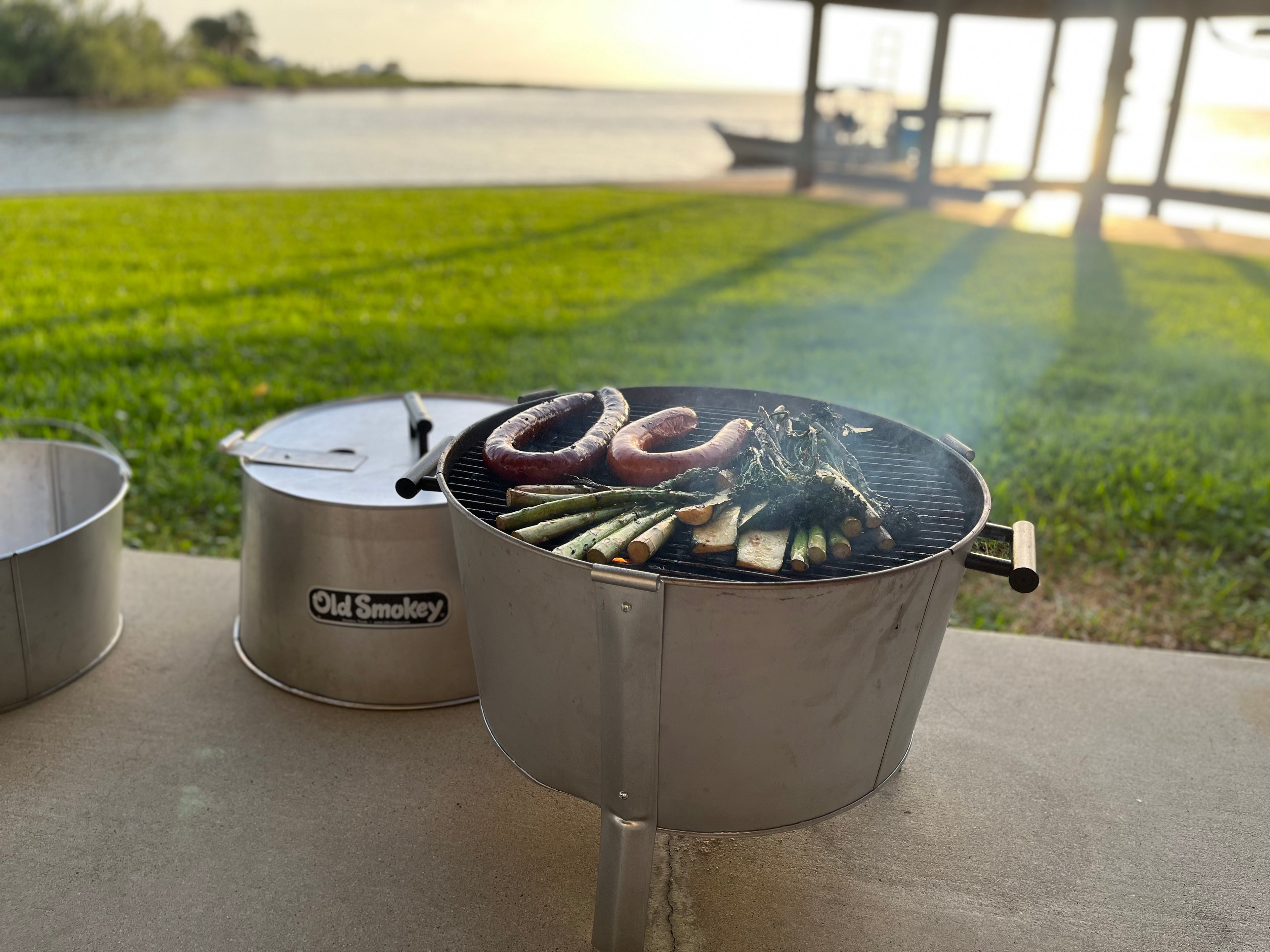 Outdoor cooking scene with a portable Old Smokey Charcoal grill by a body of water.